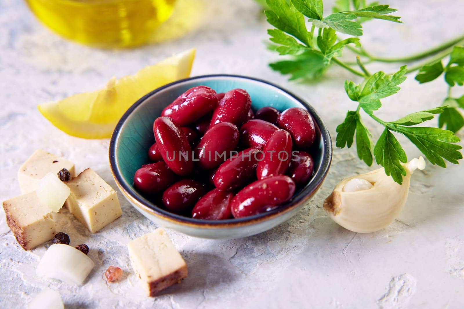 From above of delicious red kidney beans in small bowl served on table near cheese cubes garlic clove and lemon slice arranged with parsley and carafe of olive oil