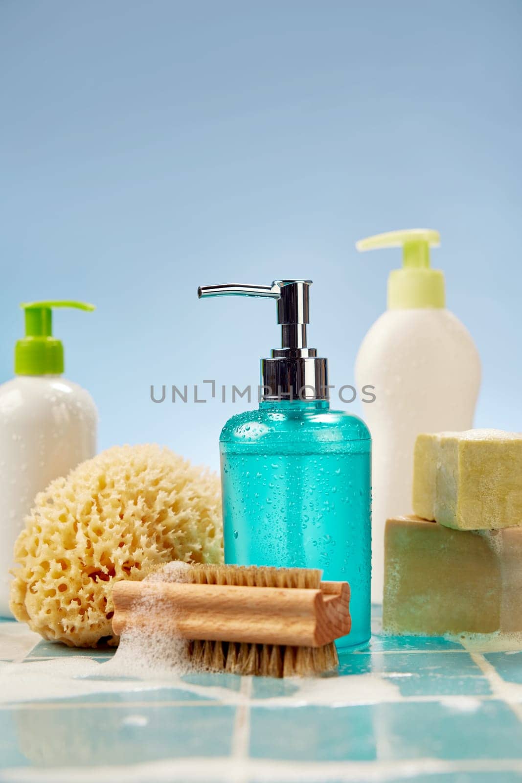 Foamy brush placed near sponge and soap on wet tiled shelf during hygienic routine against blue background