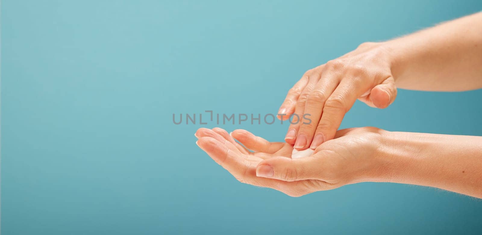 Crop female taking moisturizing cream from open palm during skin care procedure against turquoise background