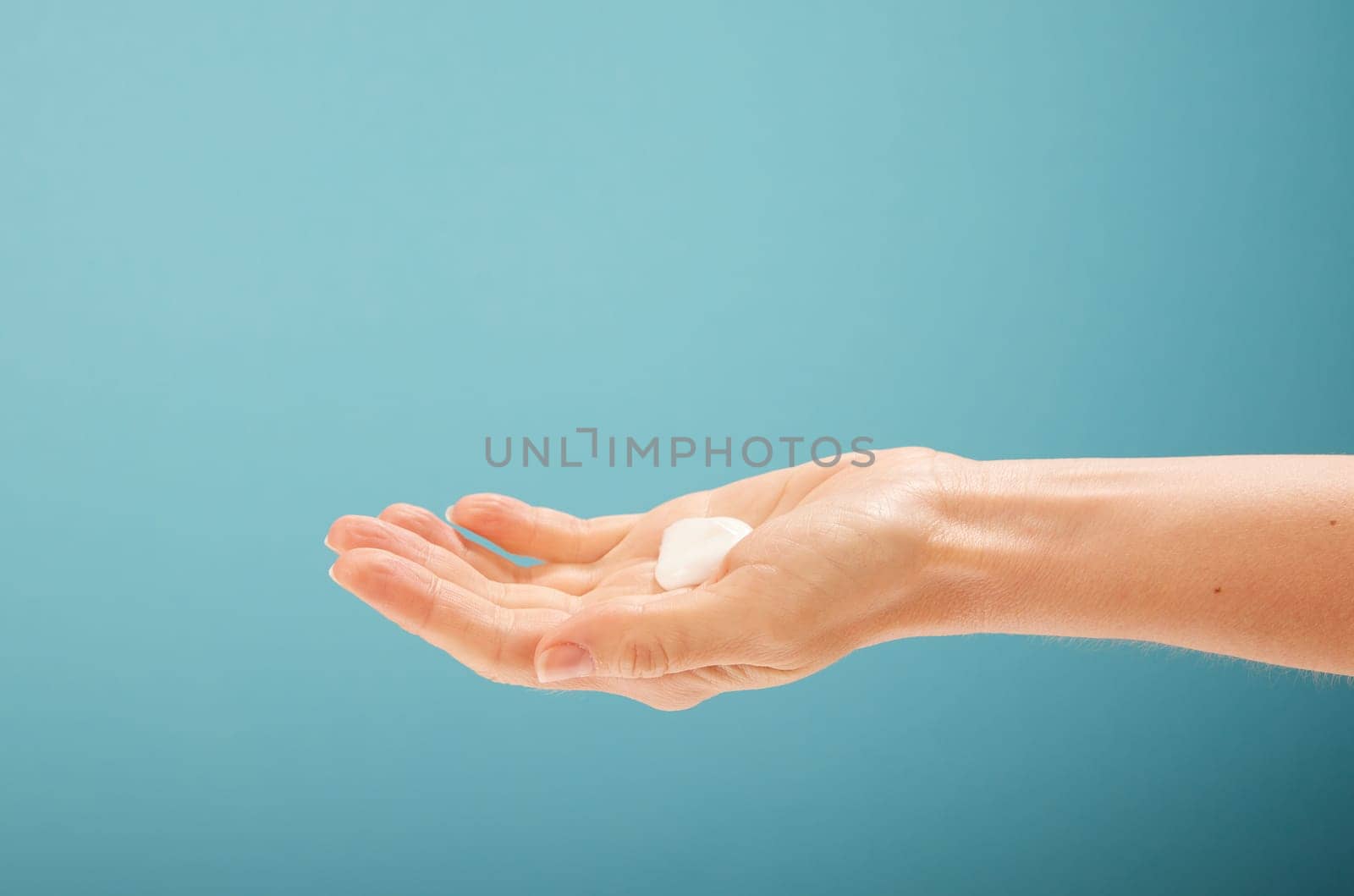 Unrecognizable woman showing open palm with drop of moisturizing cream during skin care routine against turquoise background
