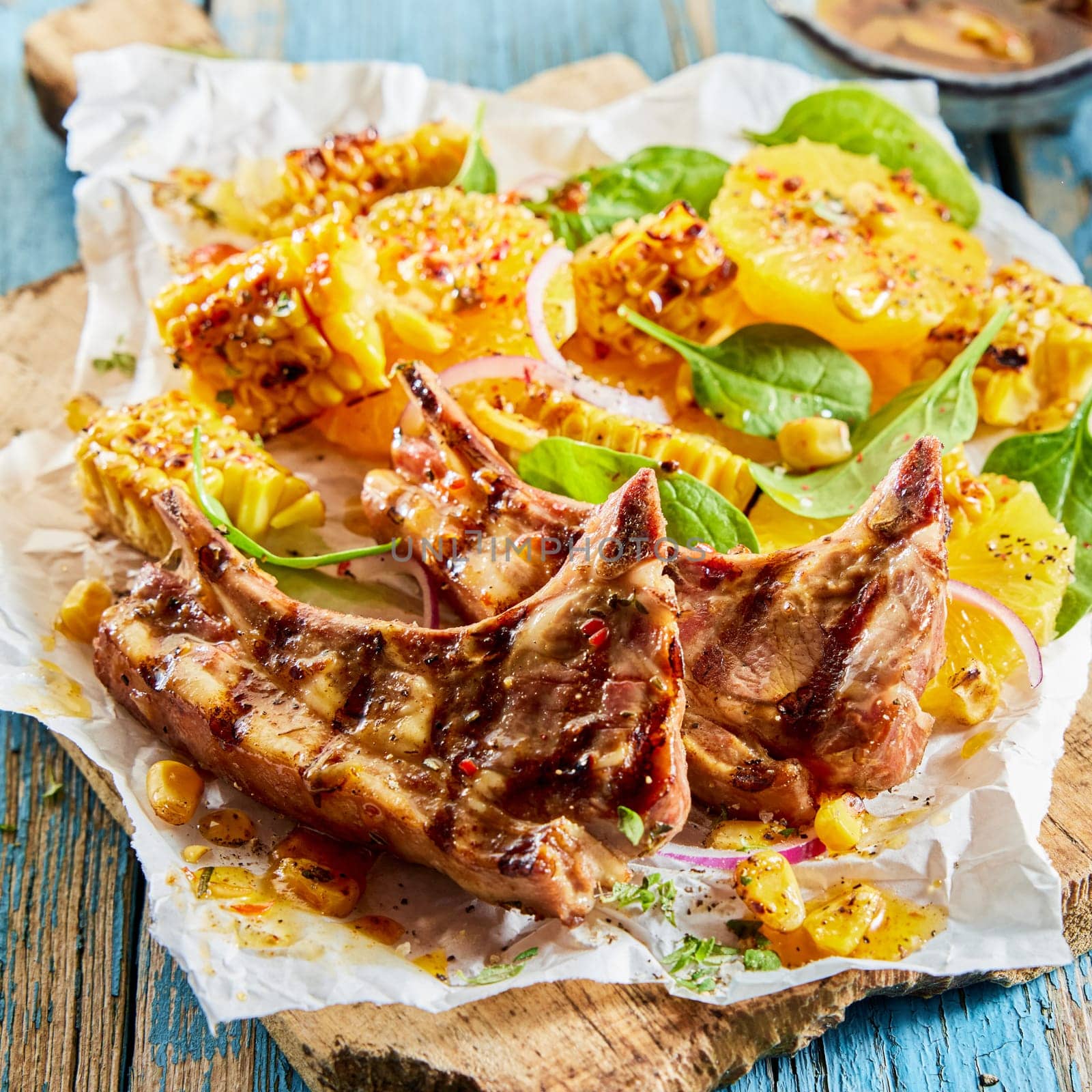 From above of appetizing grilled lamb chops with corn and green spinach leaves served with orange slices on wooden board on table
