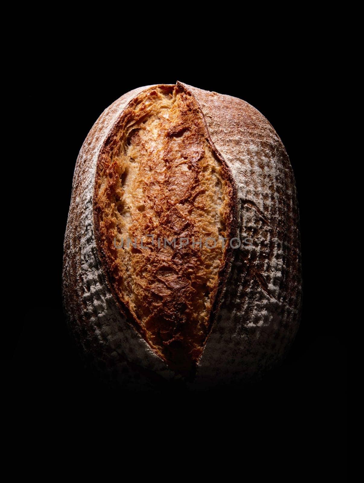 Top view of whole brown loaf of bread with crispy crust covered with flour placed on black background in dark studio