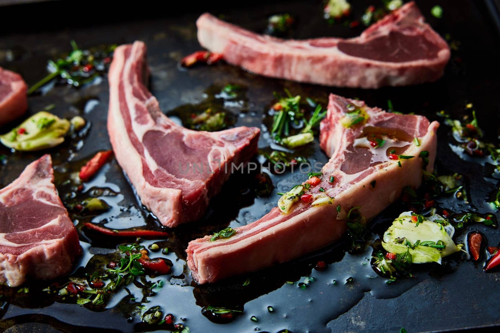Uncooked fresh lamb chops on black surface with chopped greens and chili pepper with oil in light kitchen during cooking process