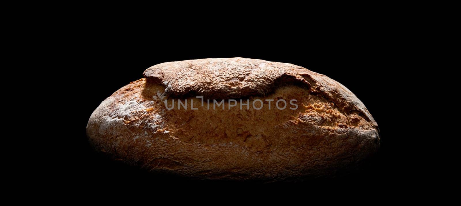 Freshly baked loaf of bread with crispy crust covered with flour placed on black background in studio with dim illumination