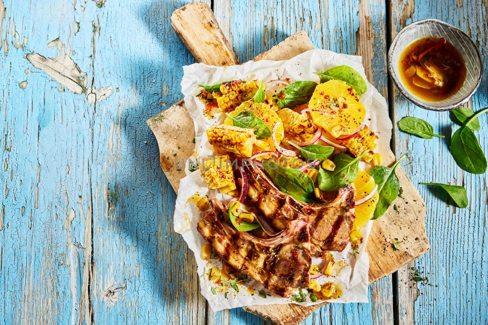 Top view of appetizing lamb chops and corn served on parchment with spinach leaves and orange slices on blue shabby table in kitchen