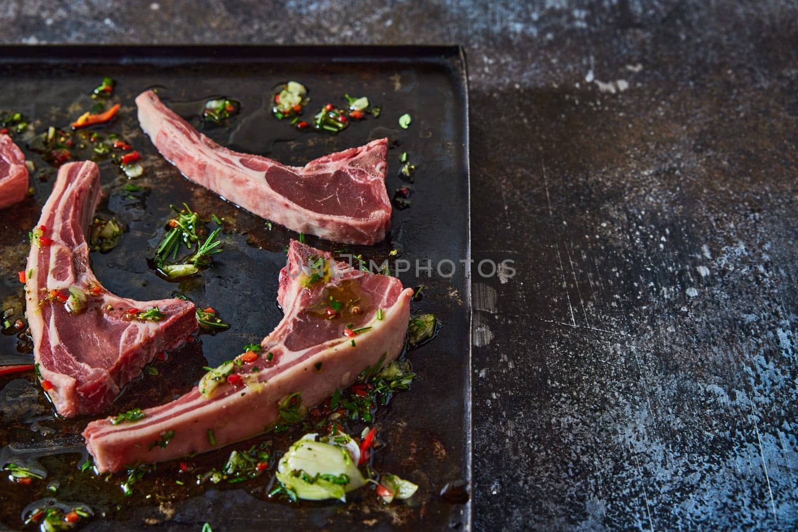From above of uncooked lamb chops placed on black tray with chopped greens and oil on dark table in light kitchen