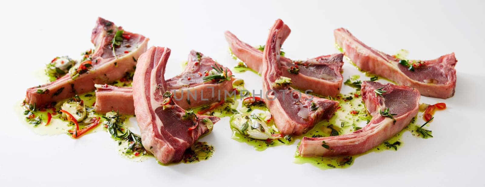 Uncooked fresh lamb chops placed on oil with chopped greens and garlic cloves on clear white background in light kitchen