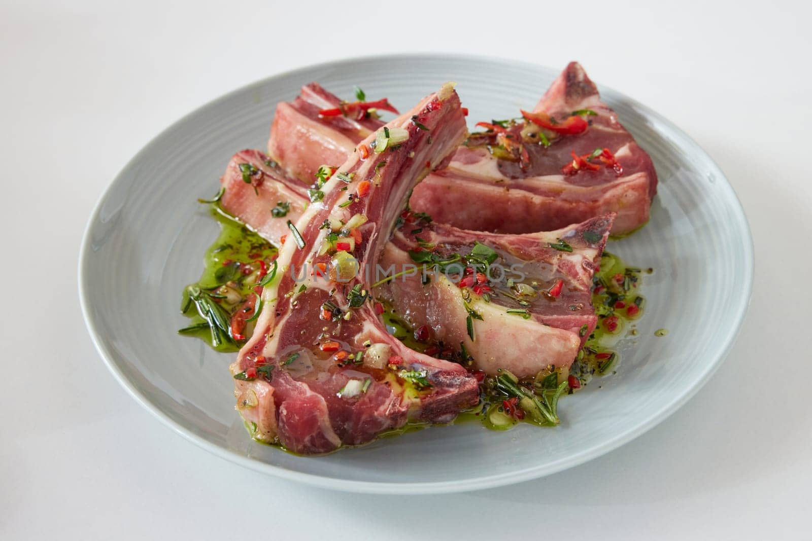 Raw lamb chops in marinate with greens and chili pepper served on ceramic plate on white background in light kitchen