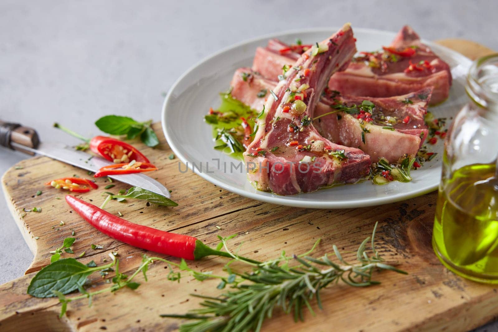 Uncooked marinated lamb chops placed on wooden chopping board with chili pepper near bottle of oil and greens in light kitchen