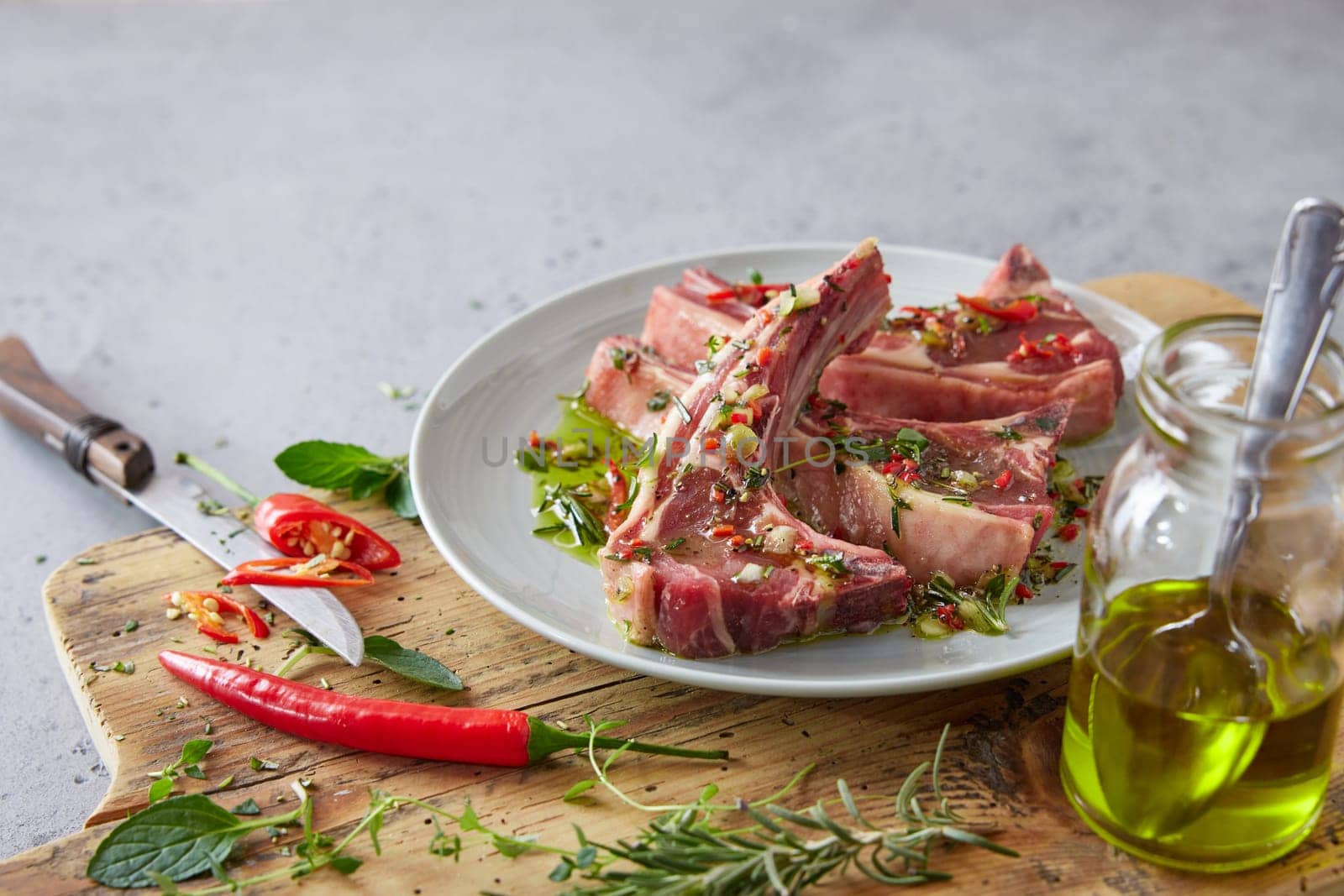 Plate with raw marinated lamb chops on wooden chopping board with oil and chili pepper on gray background in kitchen