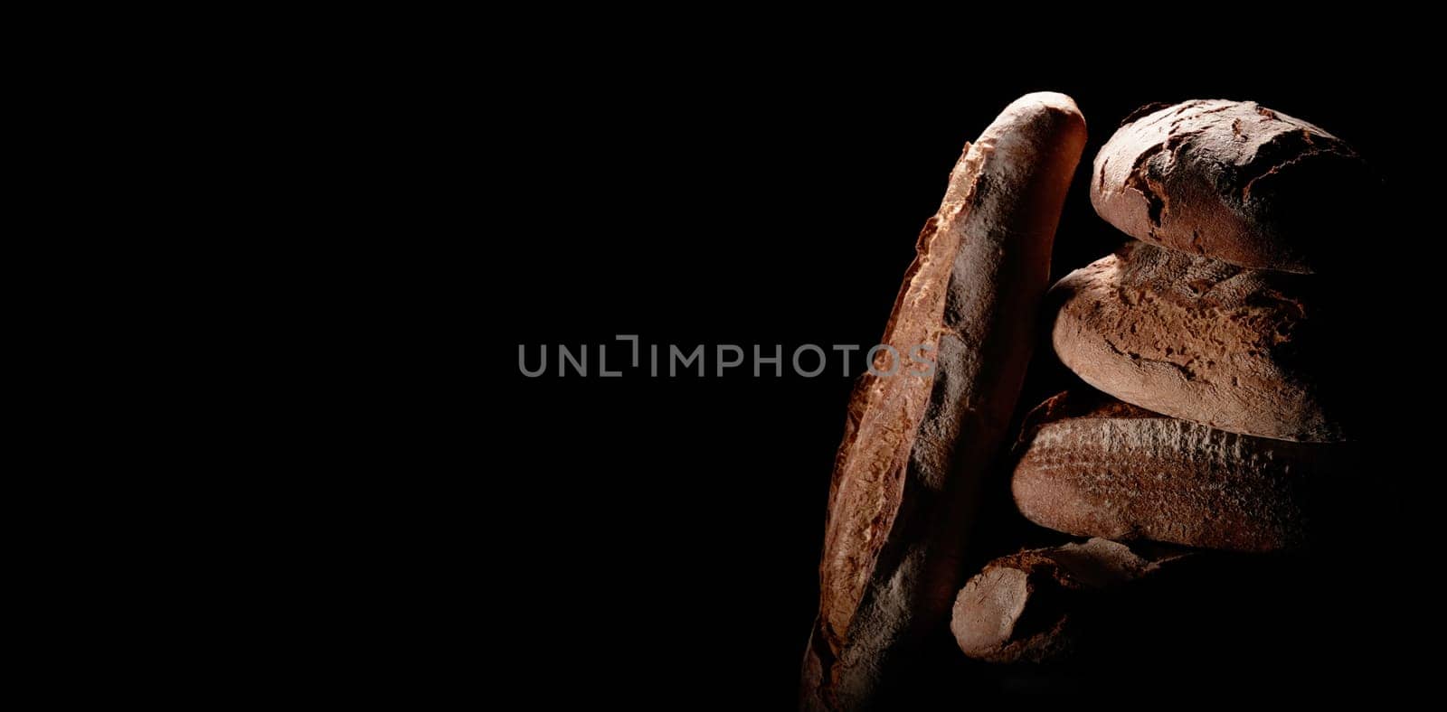 Bunch of delicious freshly baked bread loaves with baguette stacked on top of each other on black background in dark studio