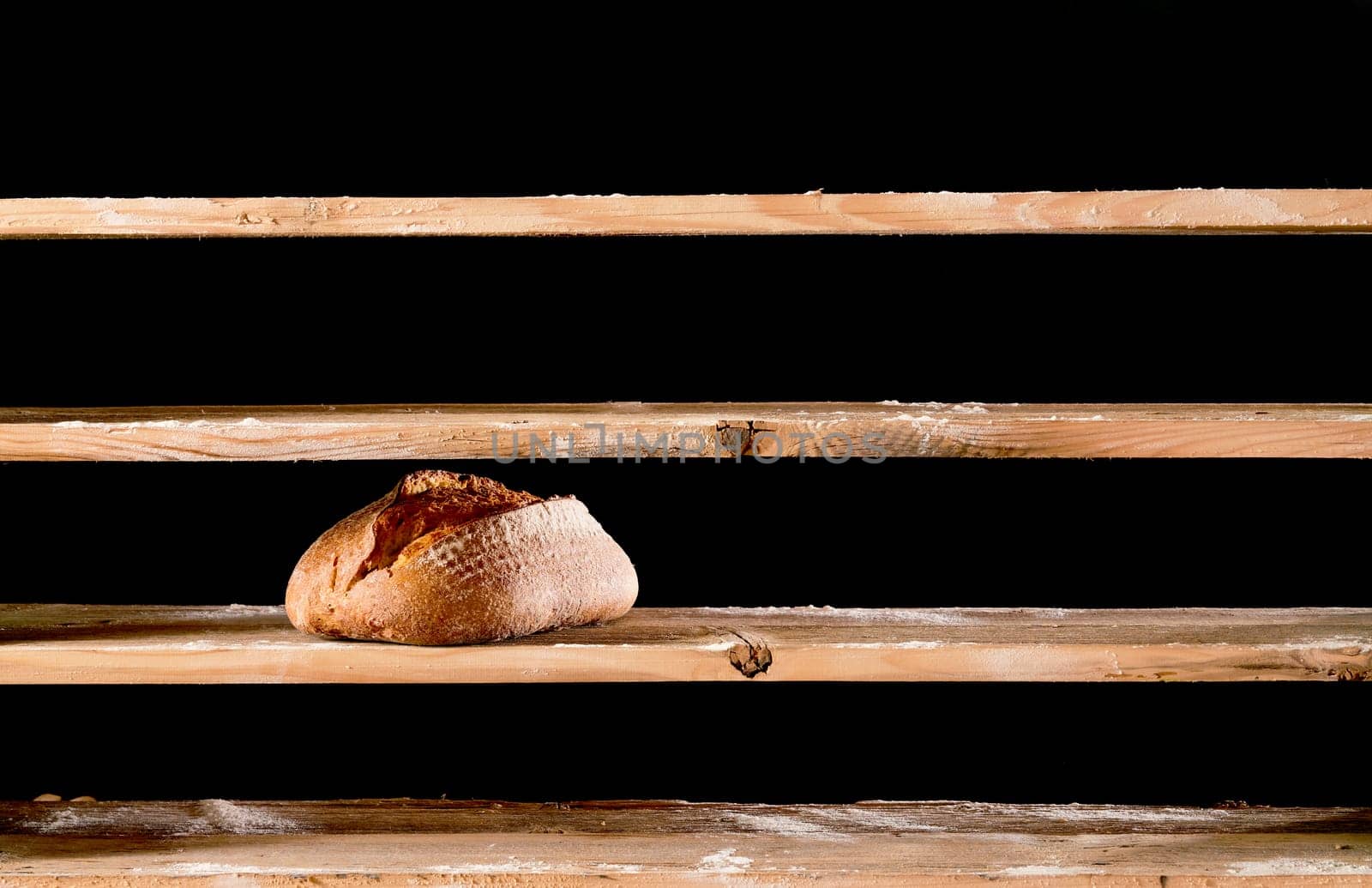One loaf of baked bread placed on empty wooden shelves with flour in light bakery on black background during scarce time