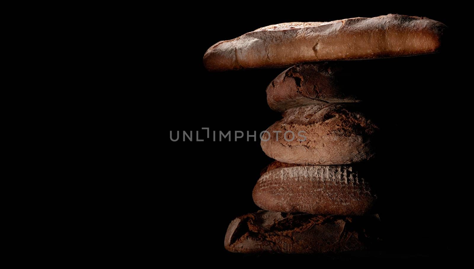 Bunch of delicious freshly baked bread loaves and baguette stacked on top of each other against black background in studio