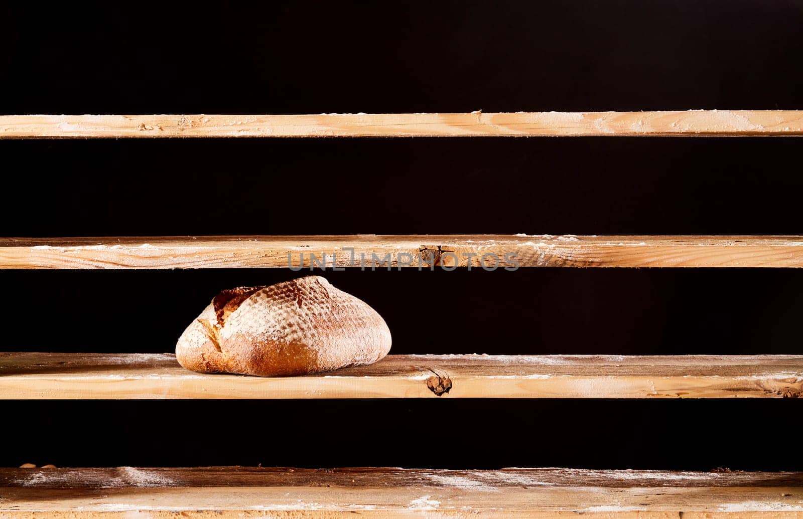 Loaf of baked bread with crust placed on empty wooden shelves in bakery on black background during scarce poverty time in bakery
