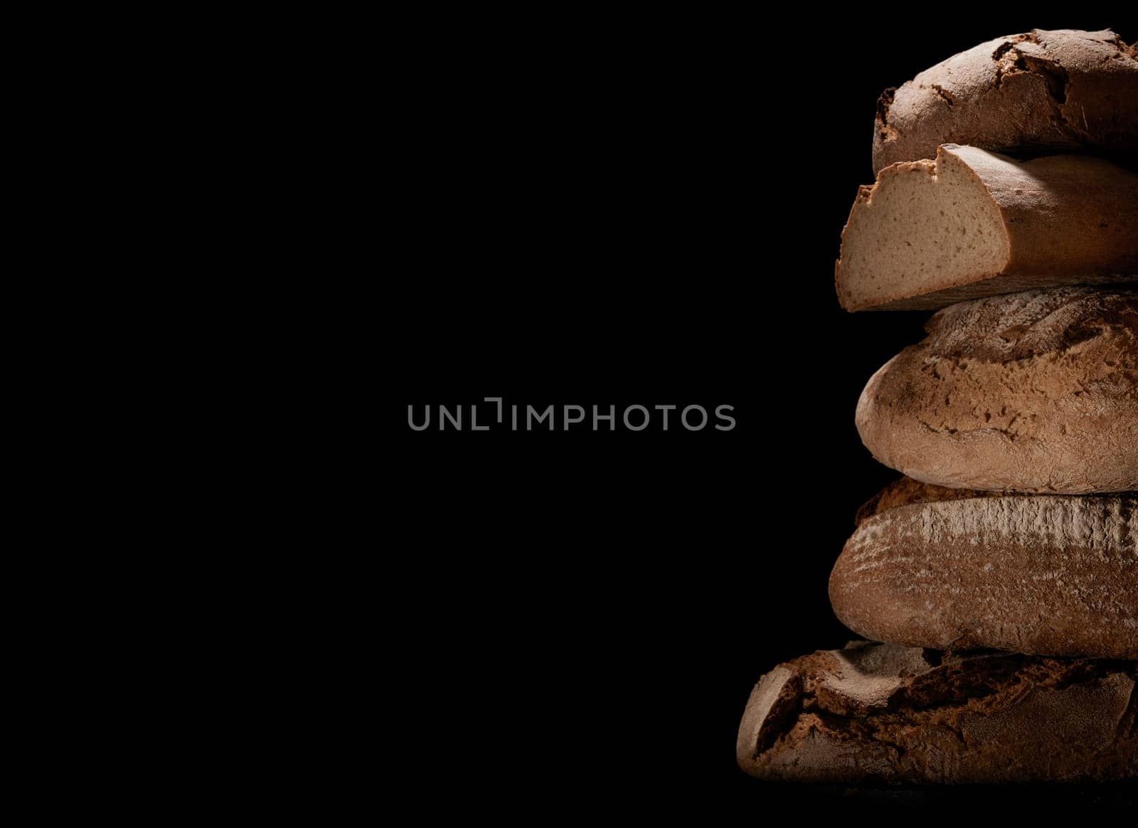 Heap of freshly baked loaves of bread with crust stacked on each other against black background in studio with obscure light
