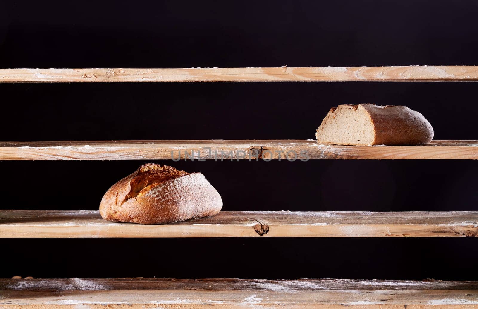 Two baked loaves of bread placed on empty wooden shelves with flour of bakehouse on black background during war time