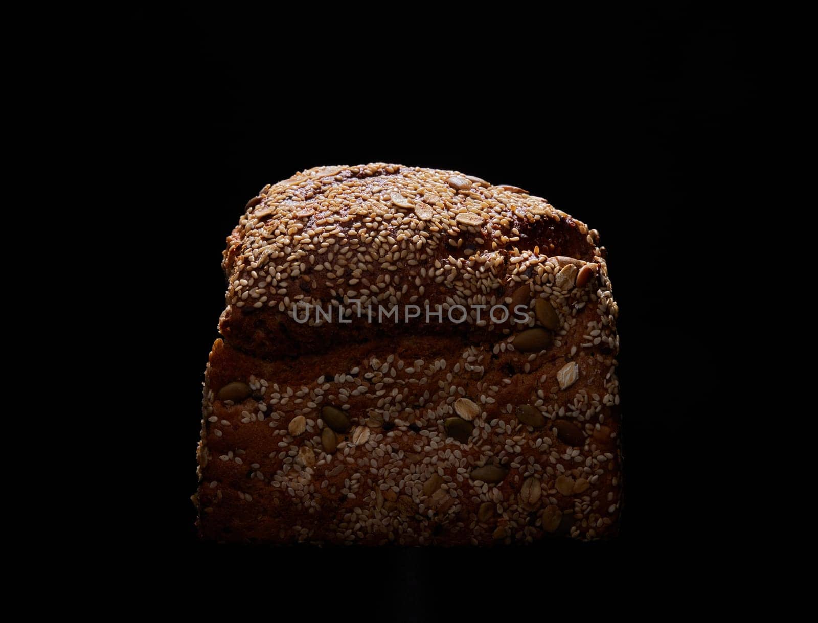 Loaf of tasty freshly baked wholegrain bread with various seeds placed on black background in dark studio with dim light