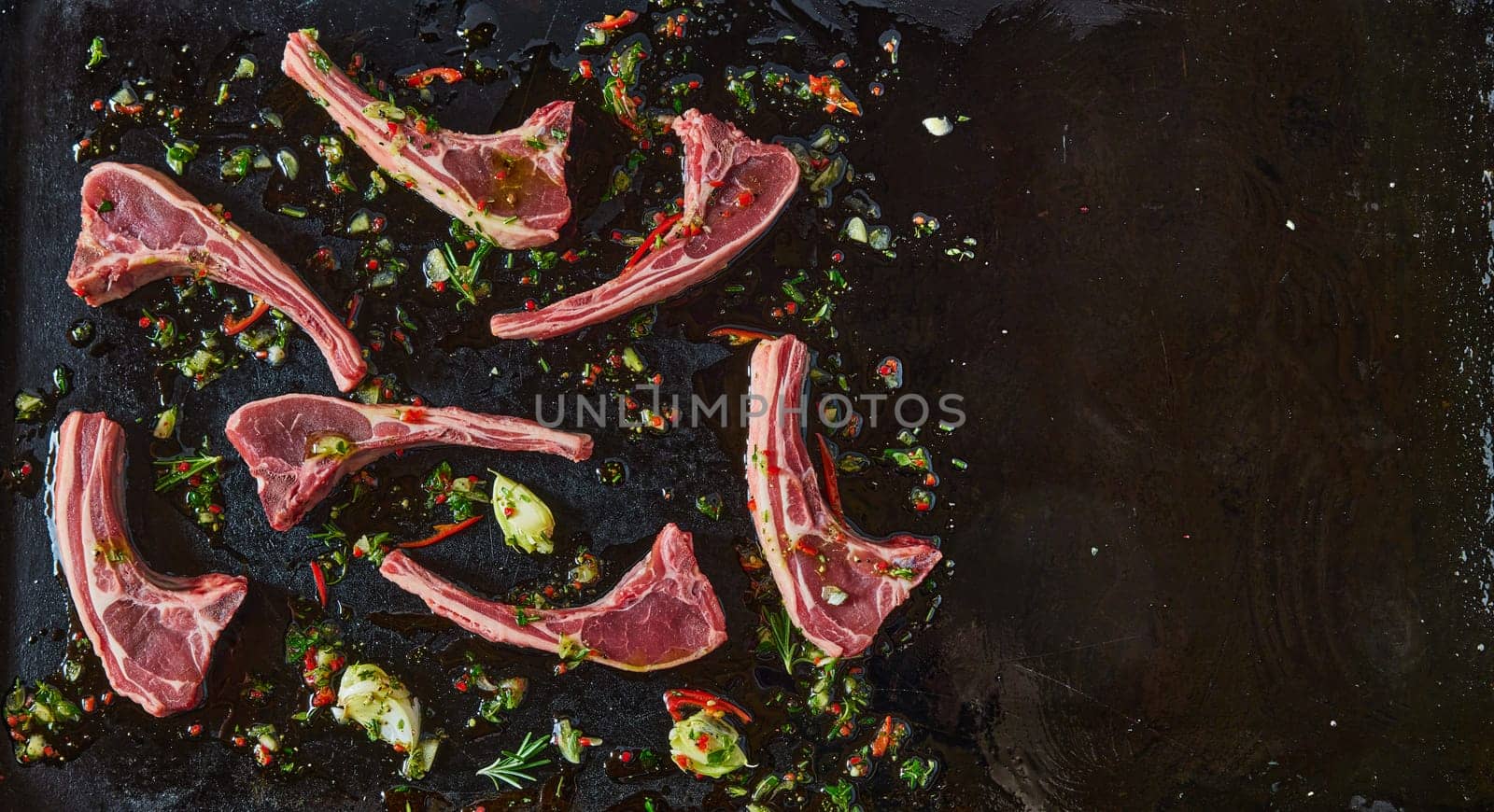 Top view of uncooked lamb chops with chili pepper on black background with chopped greens and garlic cloves in kitchen
