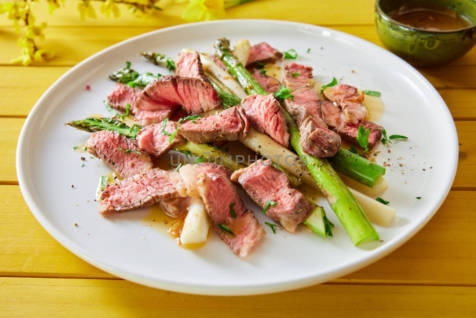 High angle of tasty beef fillet slices with asparagus and seasonings served on plate on yellow wooden table