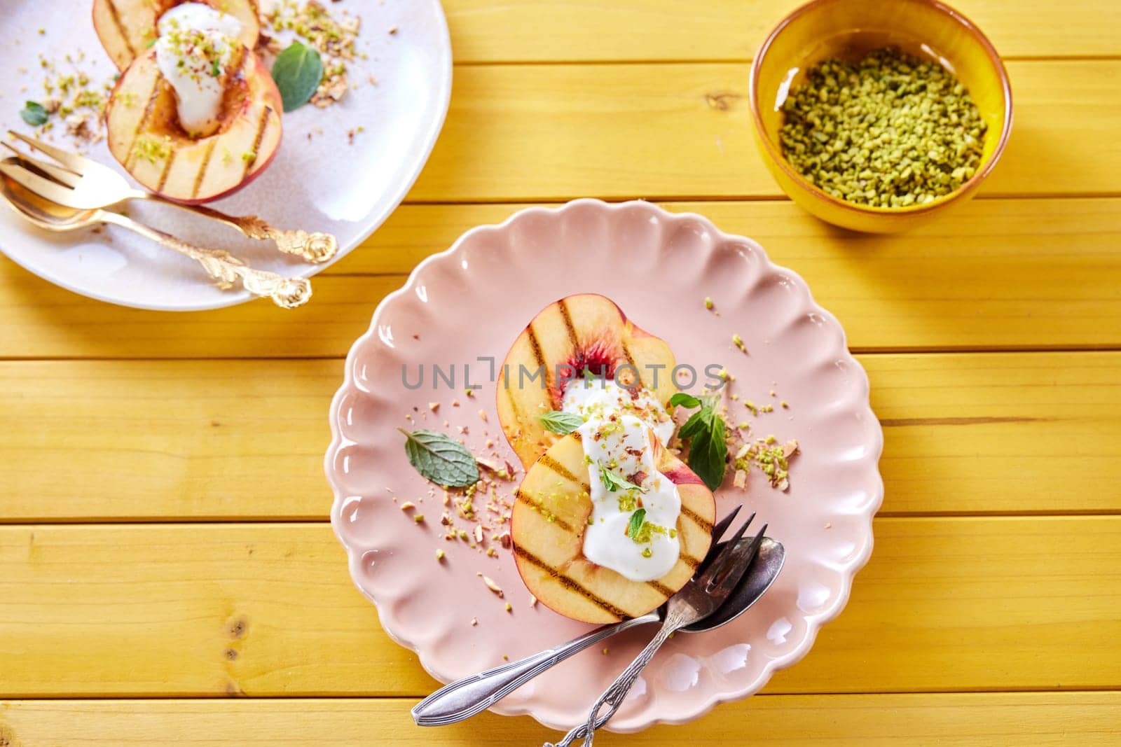 Overhead view of plate with delicious sweet nectarines with yogurt and mint leaves served on wooden table with bowl of pistachios