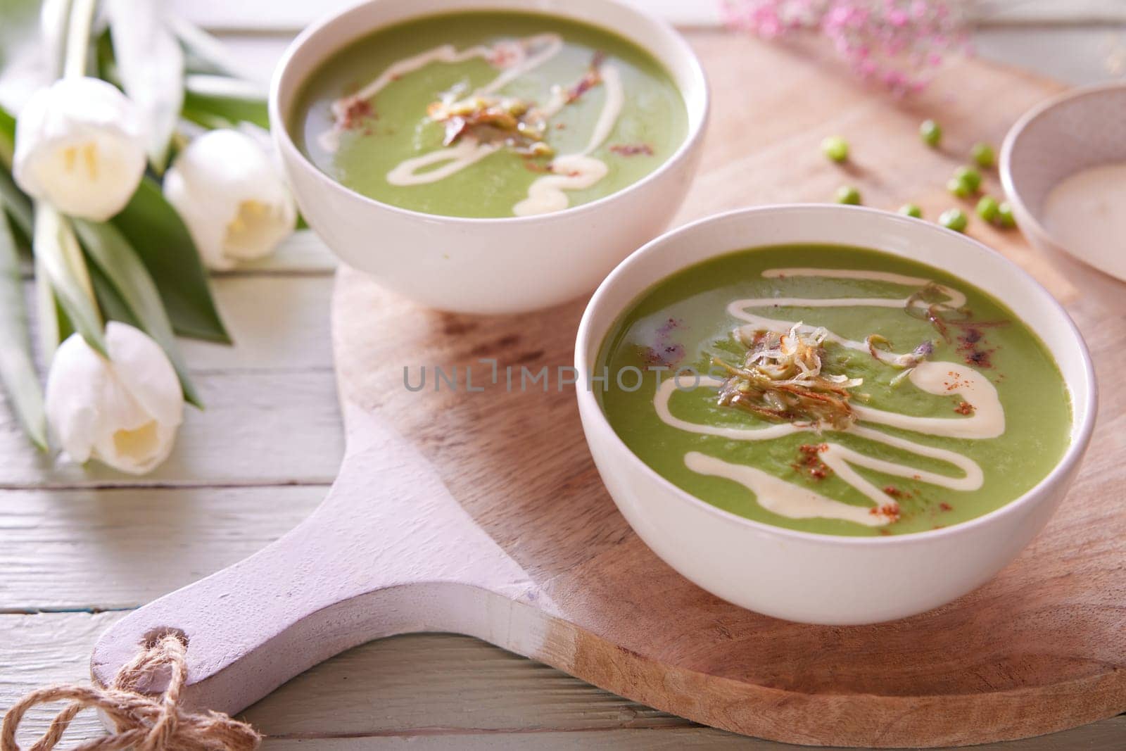 High angle of bowls with appetizing green pea soup served on wooden cutting board on table with flowers for lunch