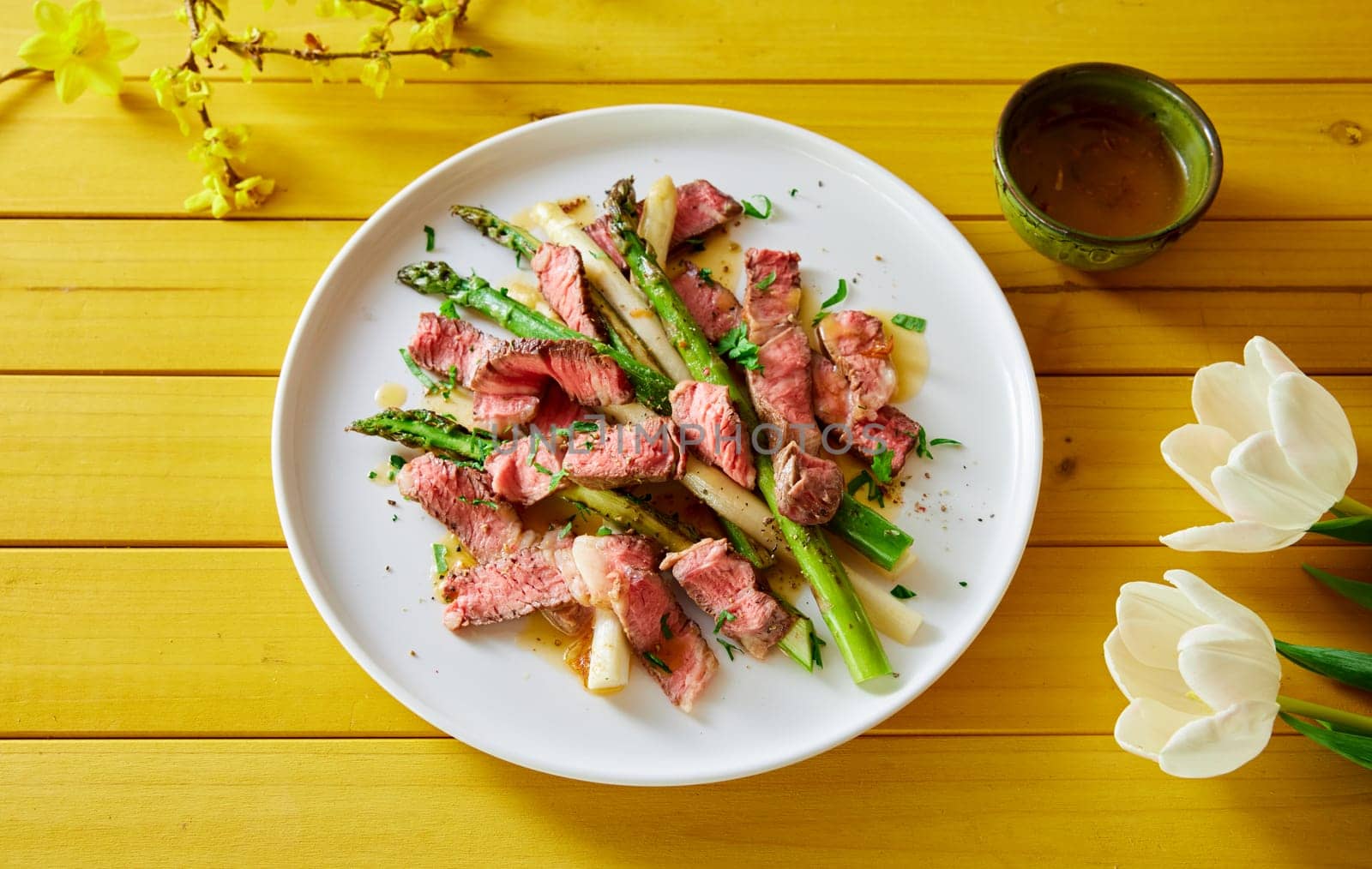Top view of plate with palatable beef fillet pieces served with asparagus on wooden table with spring flowers