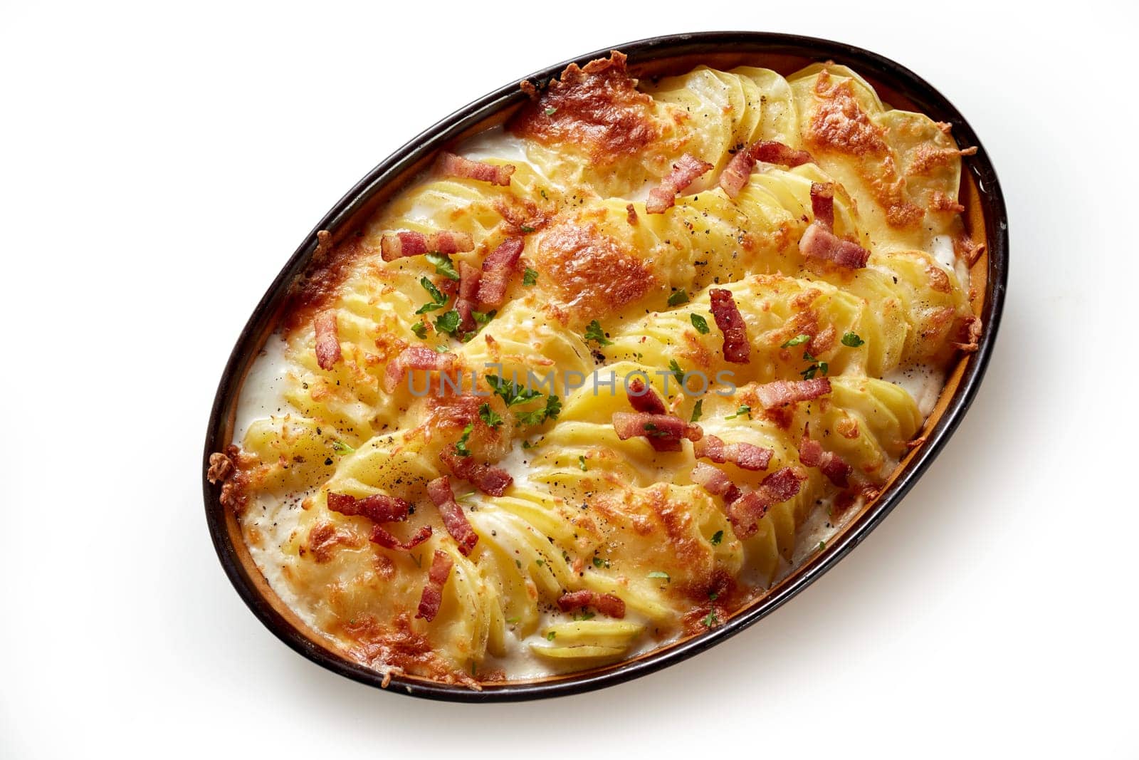 Top view of appetizing gratin with baked sliced potato and crispy bacon pieces baked in oval shaped pan placed on white background