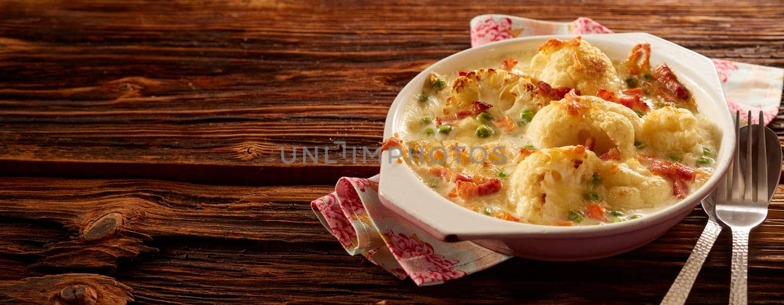 High angle of palatable baked cauliflower with cheese in bowl placed on wooden table with cutlery and napkin