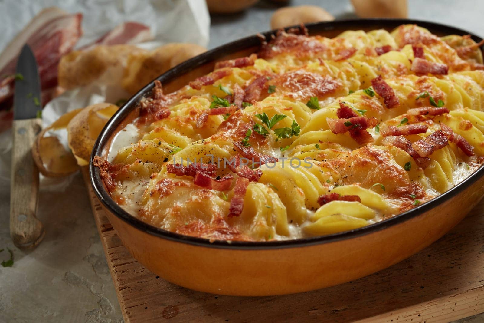 From above of yummy casserole with sliced potato bacon and cheese in oval pan placed on wooden cutting board near knife and peel in kitchen