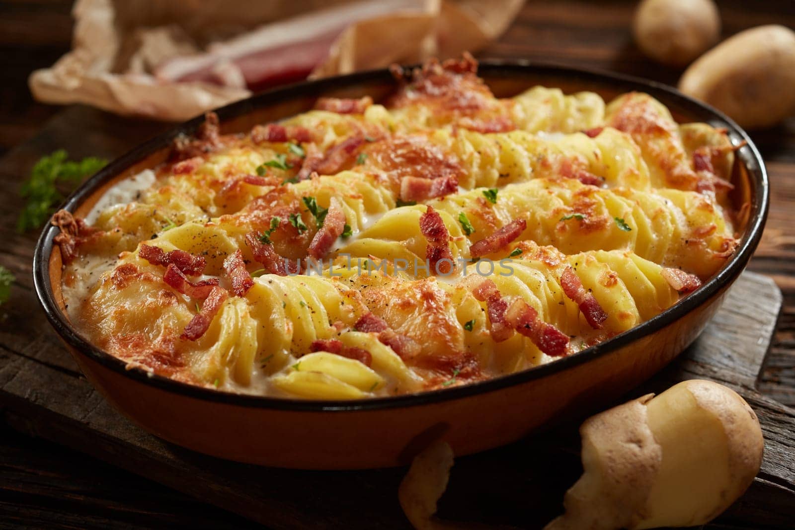 High angle of appetizing homemade dish with sliced potato and bacon baked in casserole pan and served on wooden cutting board in kitchen