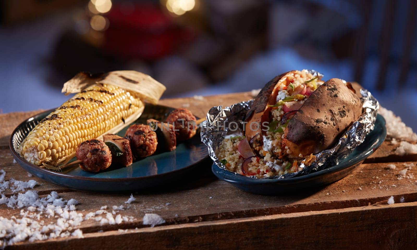 Appetizing grilled corn and meat balls on ceramic plate placed near bowl with sweet potato stuffed with bulgur on wooden table on backyard at winter night