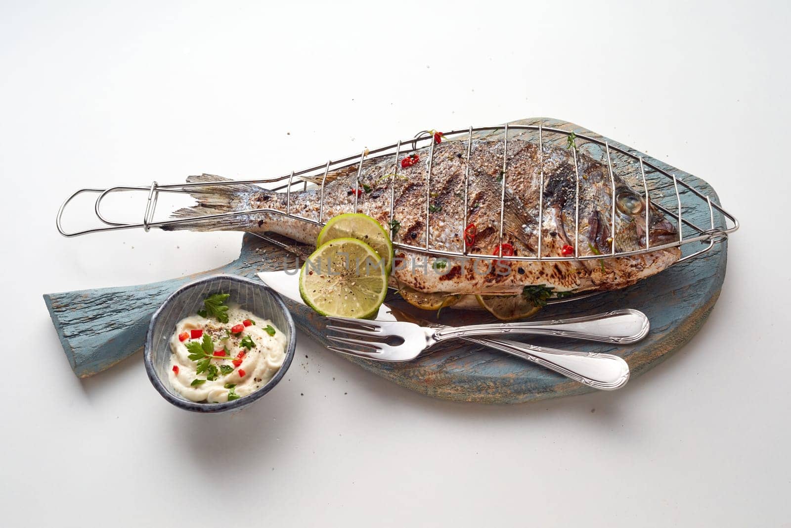 From above of delicious grilled fish served with slices of lime on wooden board placed near bowl with sauce on light table