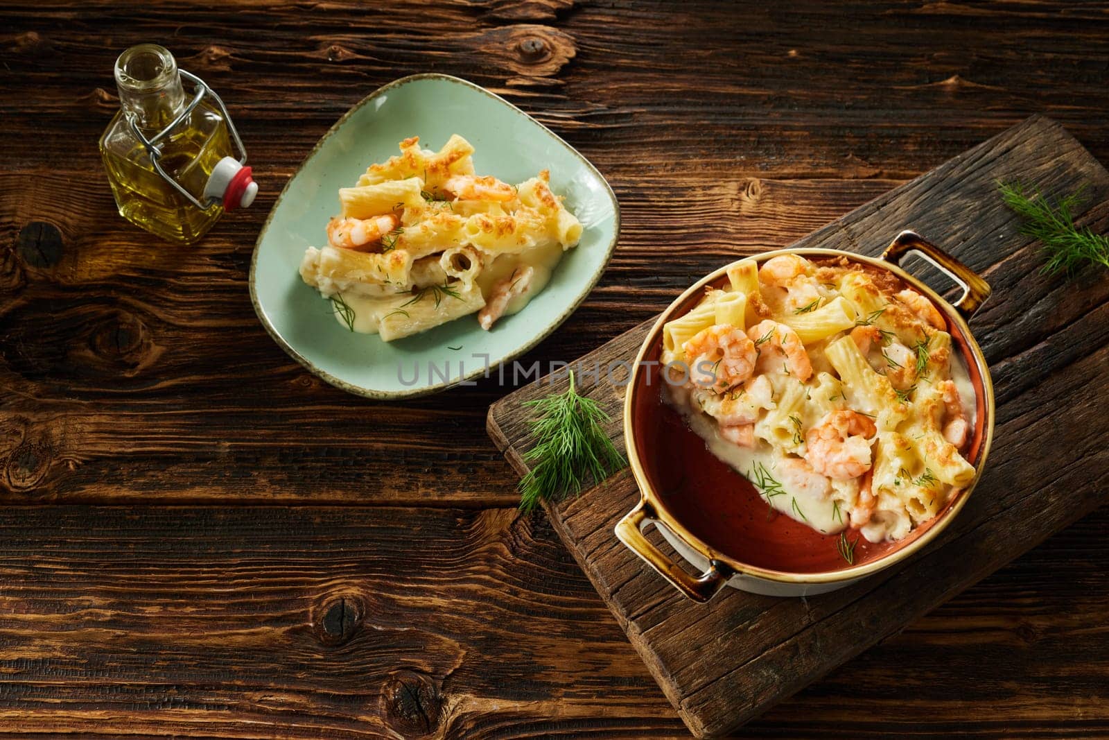 Top view of tasty homemade gratin with rigatoni pasta and shrimps served on wooden table in ceramic casserole pan and plate with bottle of olive oil