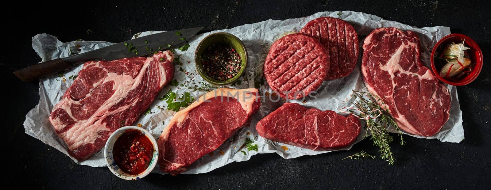 Top view of raw sirloin and ribeye steaks served with burger patties and cups of sauces and condiments on creased paper near knife on black background