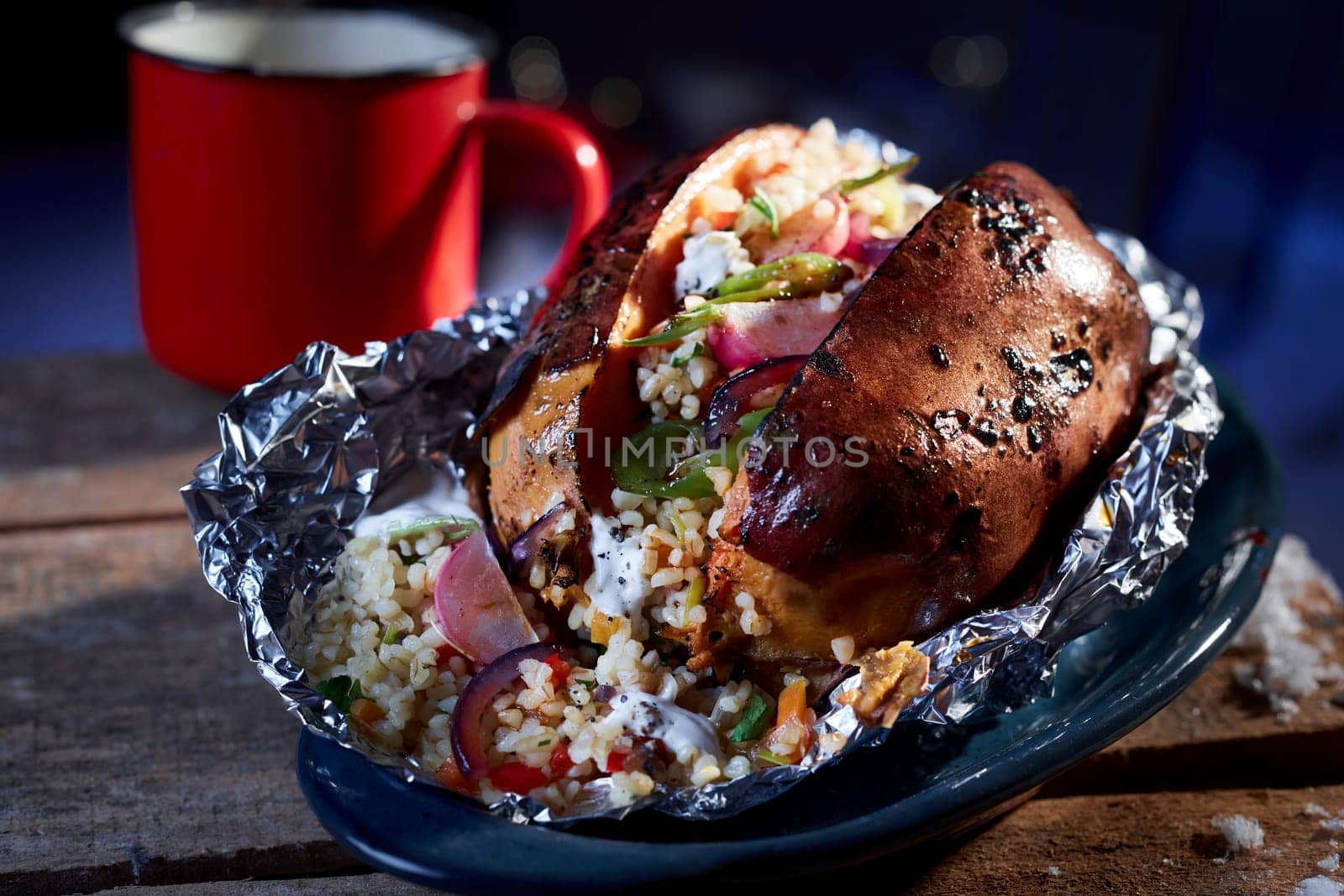 Tasty vegetarian grilled sweet potato served cooked with bulgur and vegetables in foil in ceramic bowl placed on wooden table at winter night