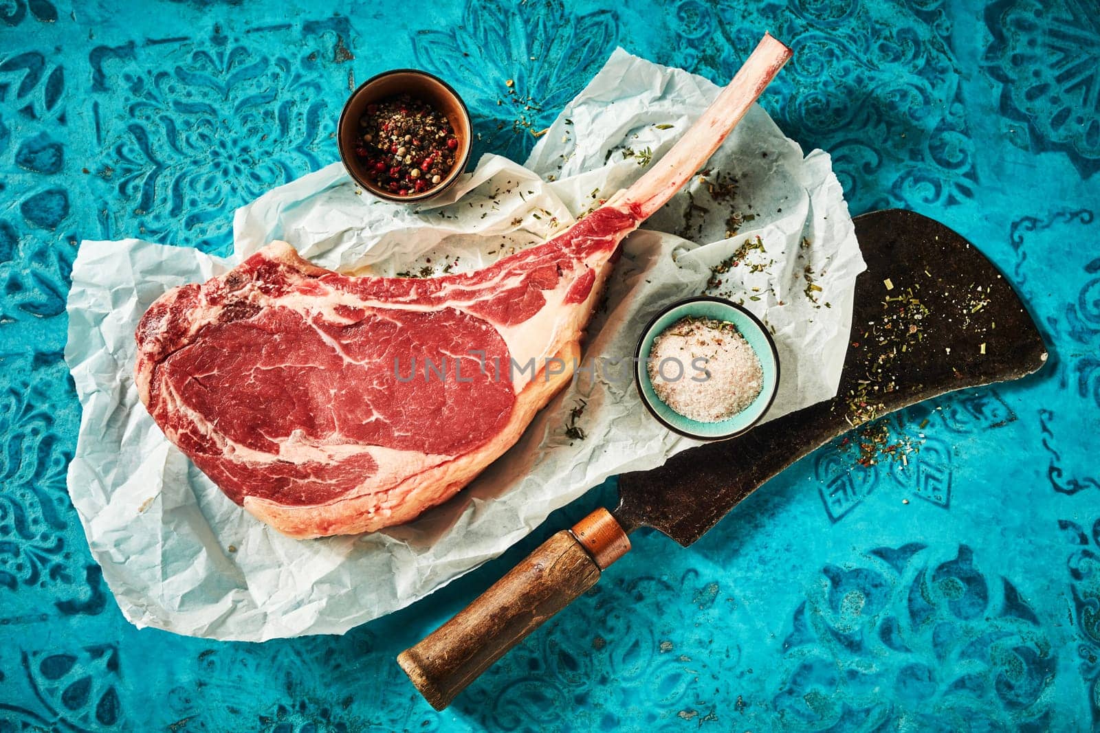 Top view of ribeye served on long rib with cup of salt and pepper on paper and chopping ax on blue table