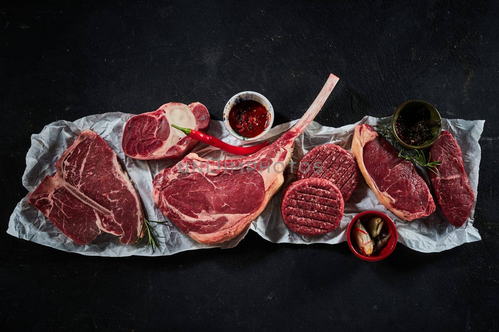 Top view of assorted raw beef steaks including t bone tomahawk sirloin served with different spices and chili pepper and rosemary twigs