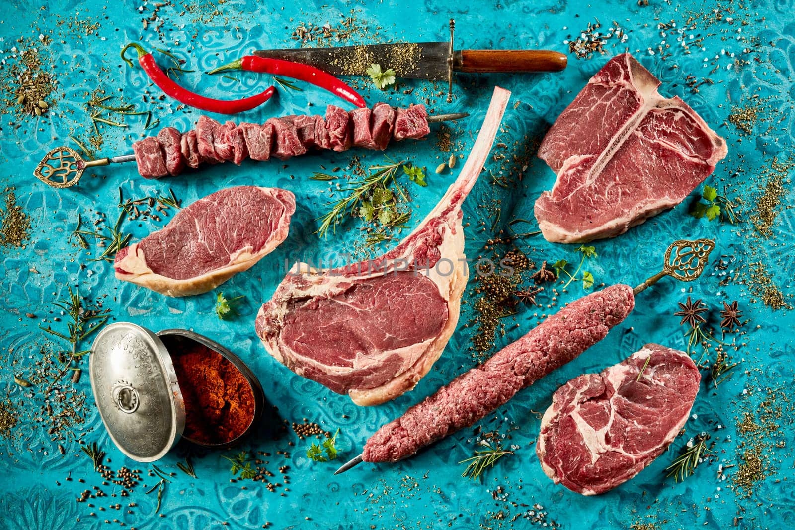 Top view of set of various types of meat steaks placed among rosemary sprigs and seasonings on blue table