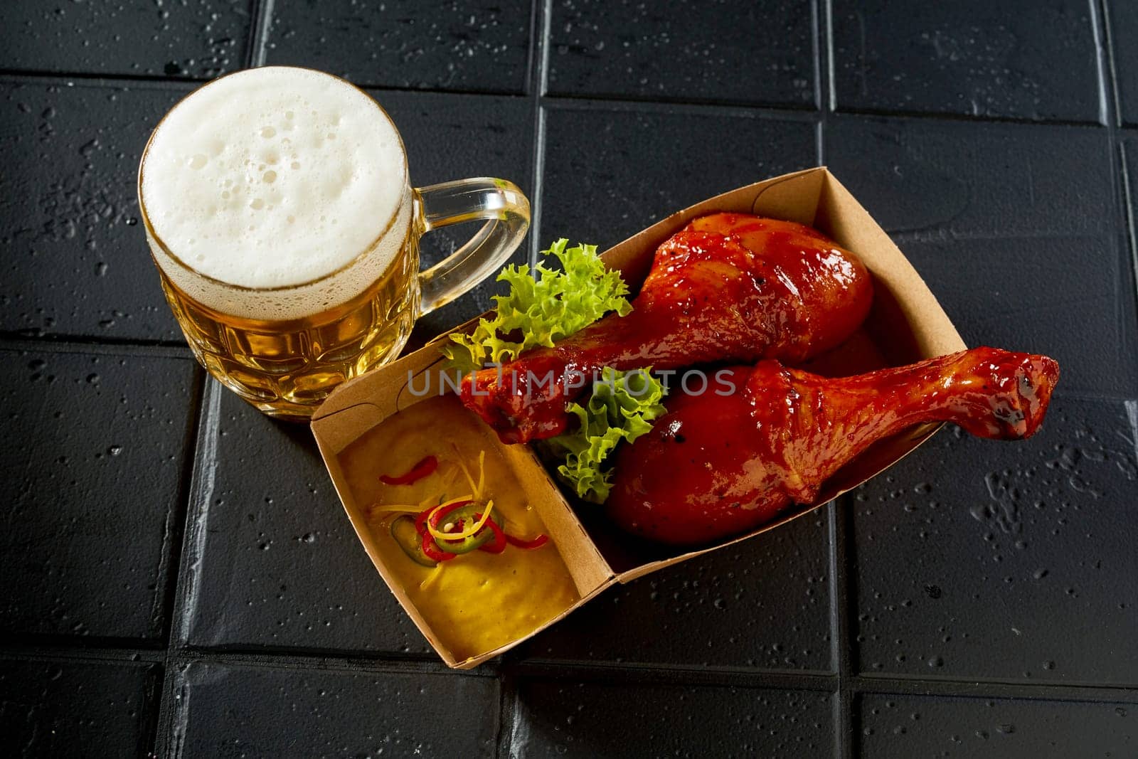 From above of appetizing chicken drumsticks served with lettuce leaf and sauce placed near glass of beer on black table in cafe