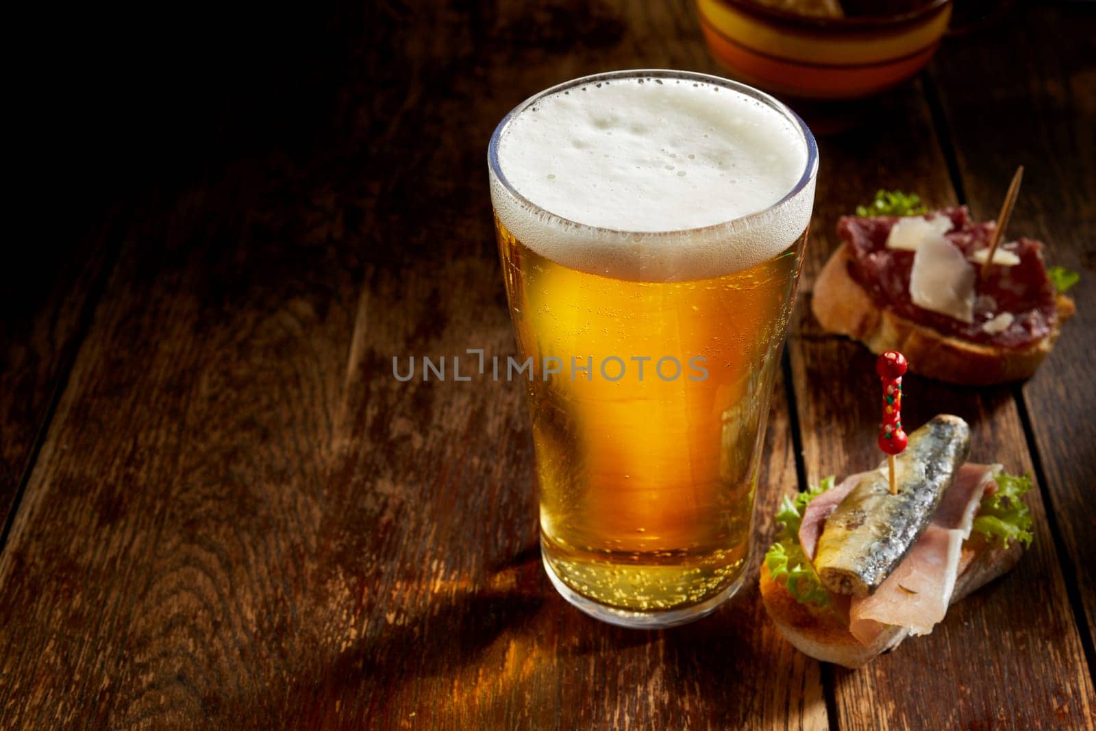 Pint of cold draft beer in a tall glass with delicious assorted tapas on fresh baguette served on a rustic counter with copyspace in a pub or tavern