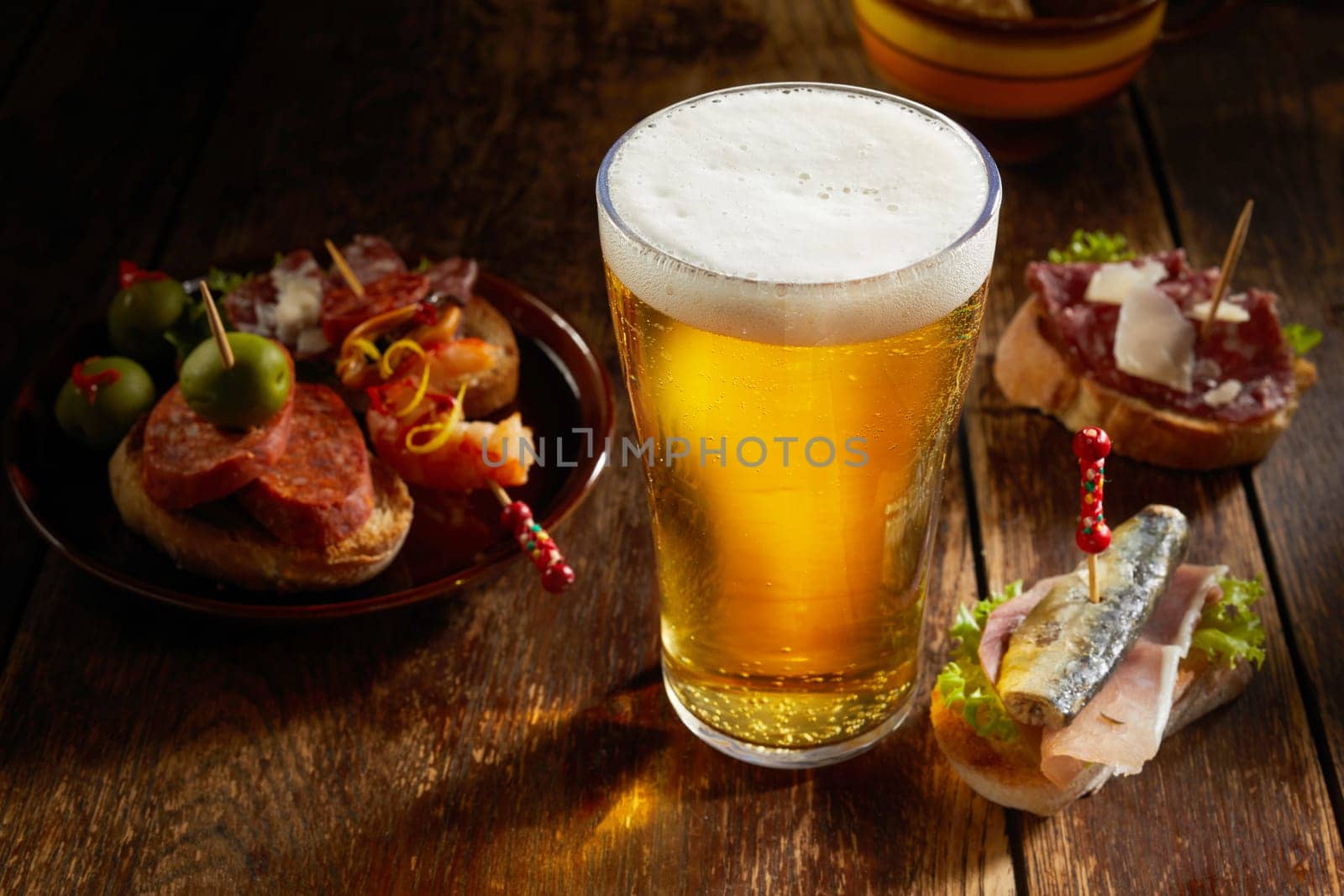 Tasty selection of savory Spanish tapas served with a cold pint of beer with frothy head on a rustic shadowy wooden counter in a tavern in a high angle close up view