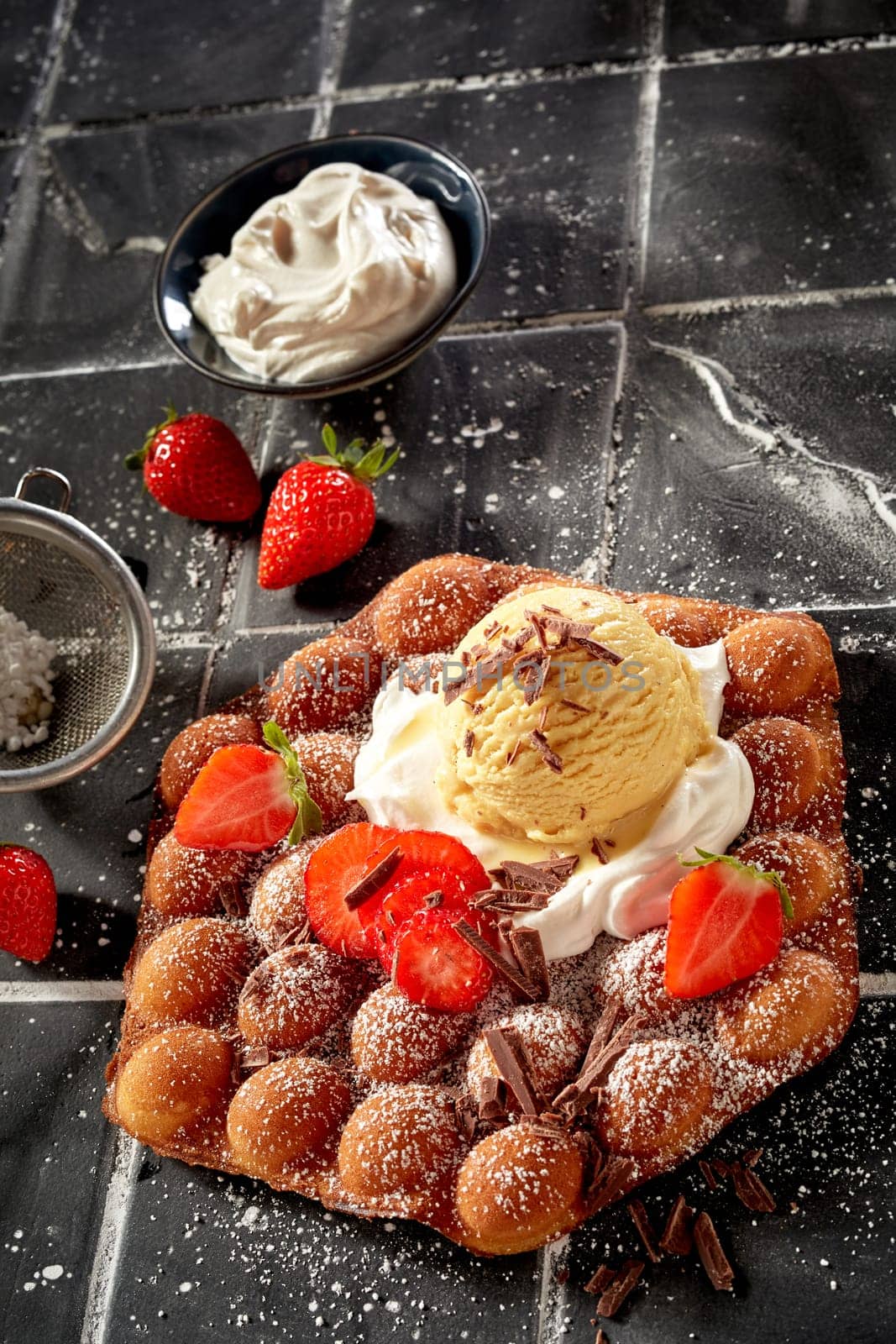 Delicious bubble waffle with ripe strawberries and whipped cream topped with vanilla ice cream drizzled with chocolate flakes and icing sugar for a gourmet summer dessert