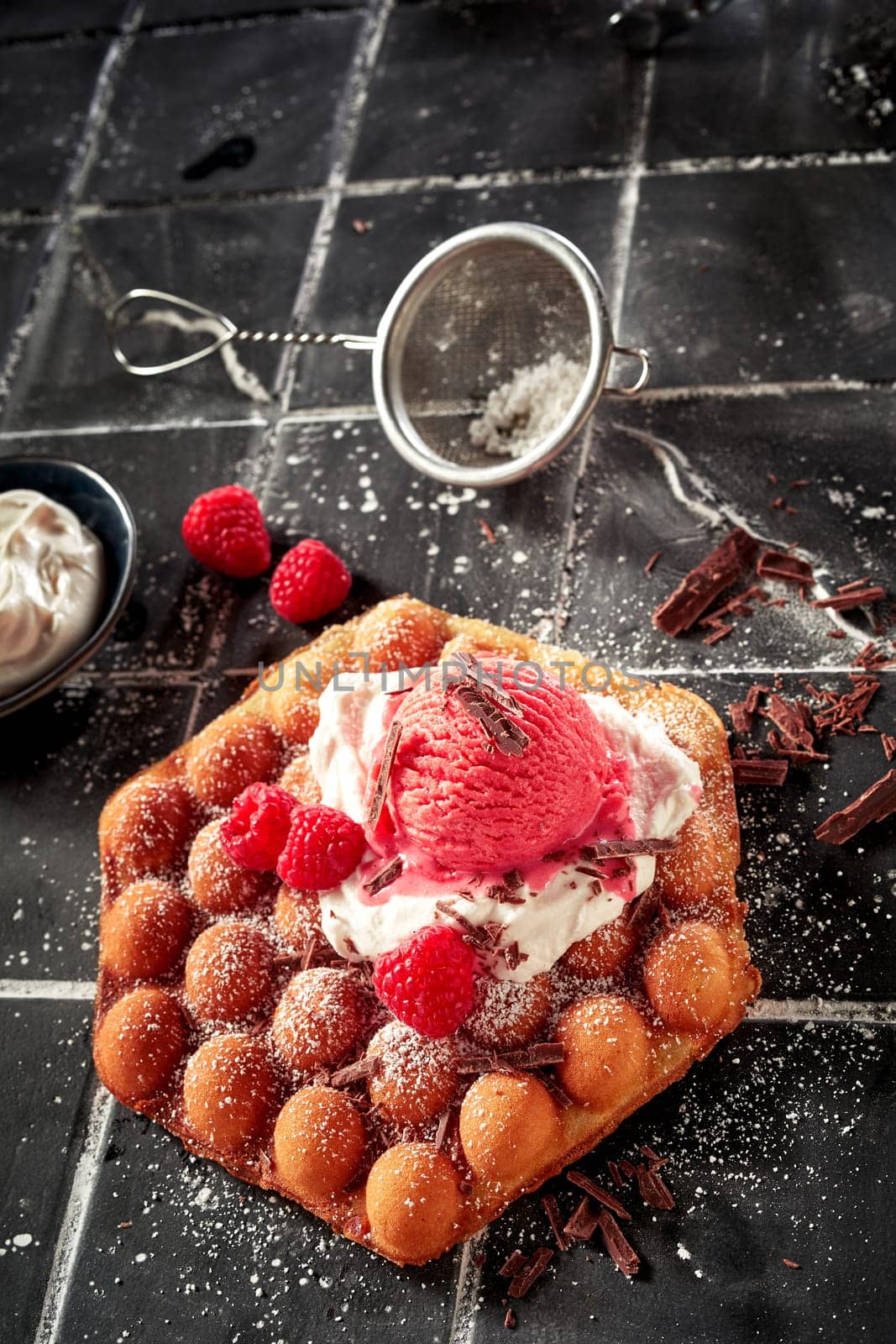 Tasty fresh raspberry and bubble waffle summer dessert with berry ice-cream on a bed of whipped cream sprinkled with chocolate flakes in a high angle vertical view on tiles with copyspace