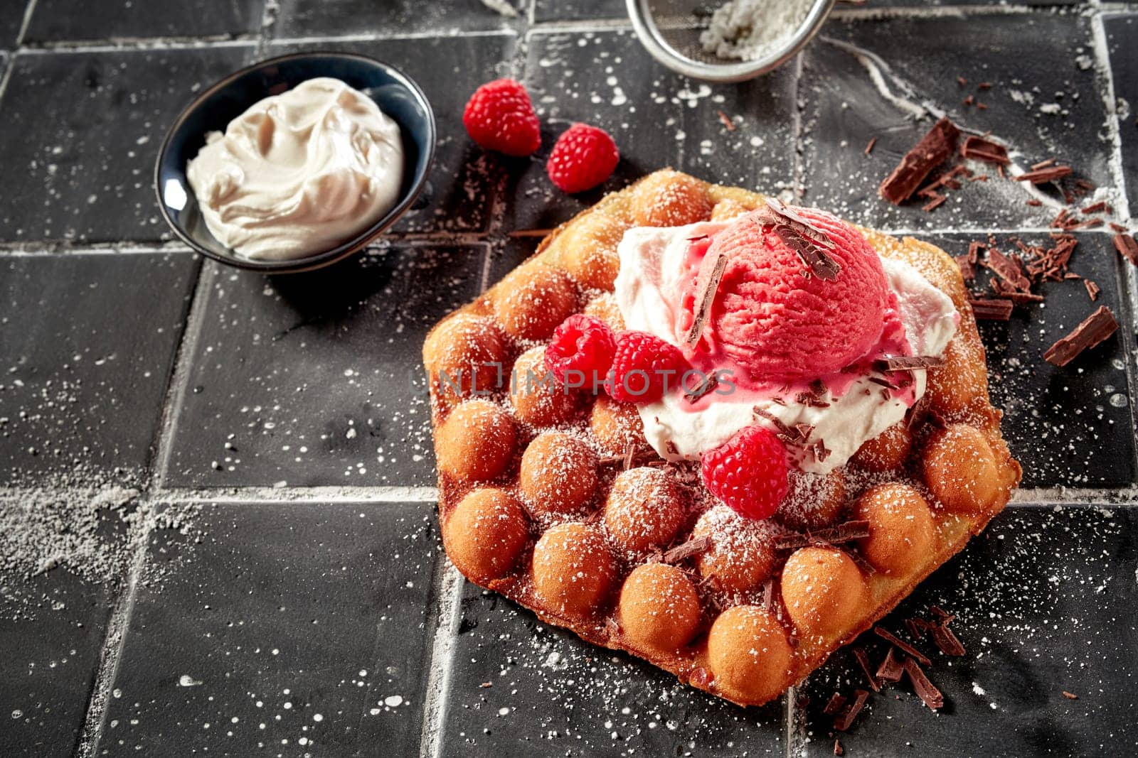 Hexagonal crispy golden bubble waffle or egg waffle topped with whipped cream, berry ice-cream and fresh tropical raspberries sprinkled with flaked chocolate in high angle with copyspace for menu advertising