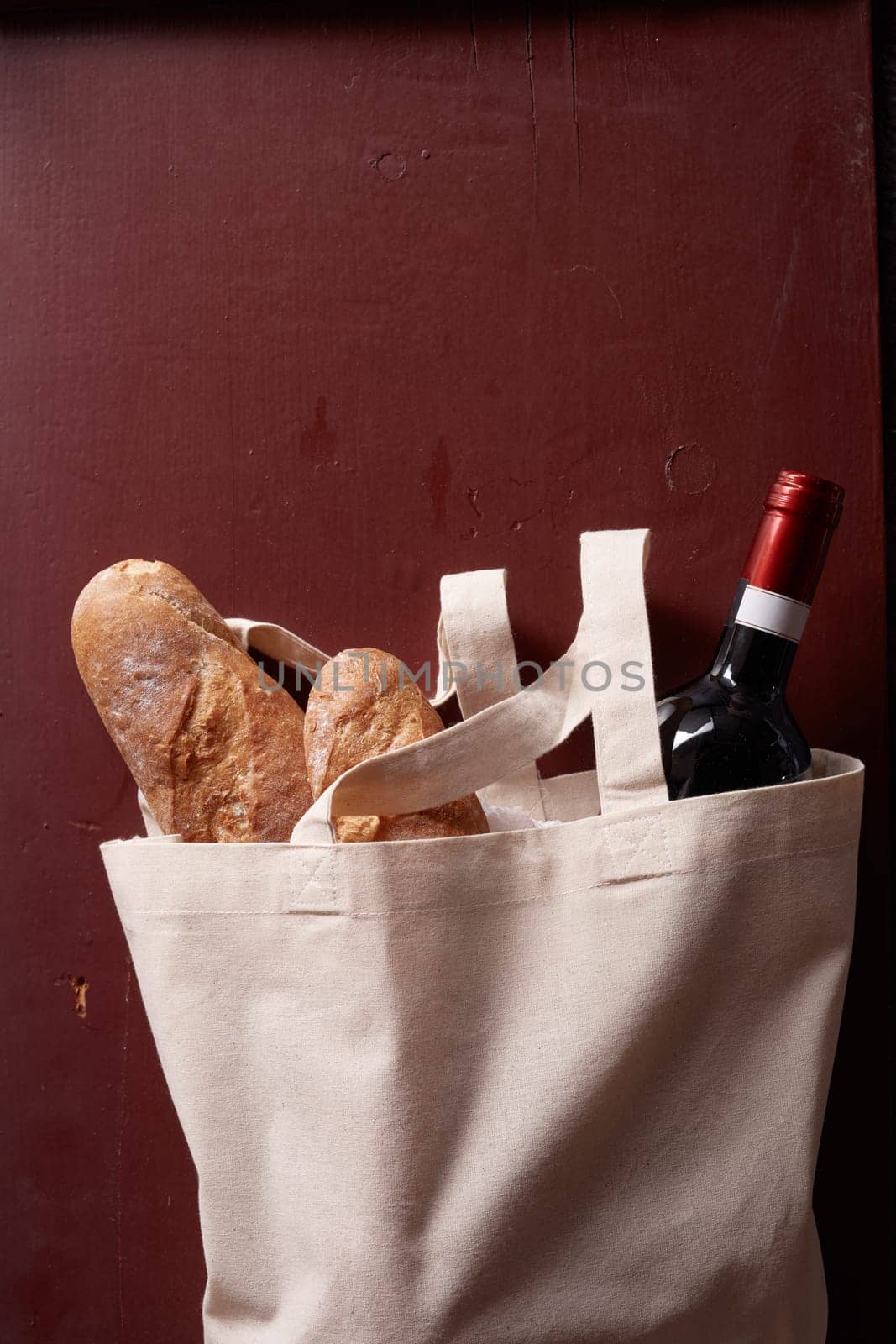 From above of bottle of wine and fresh baguettes in textile bag placed on red background