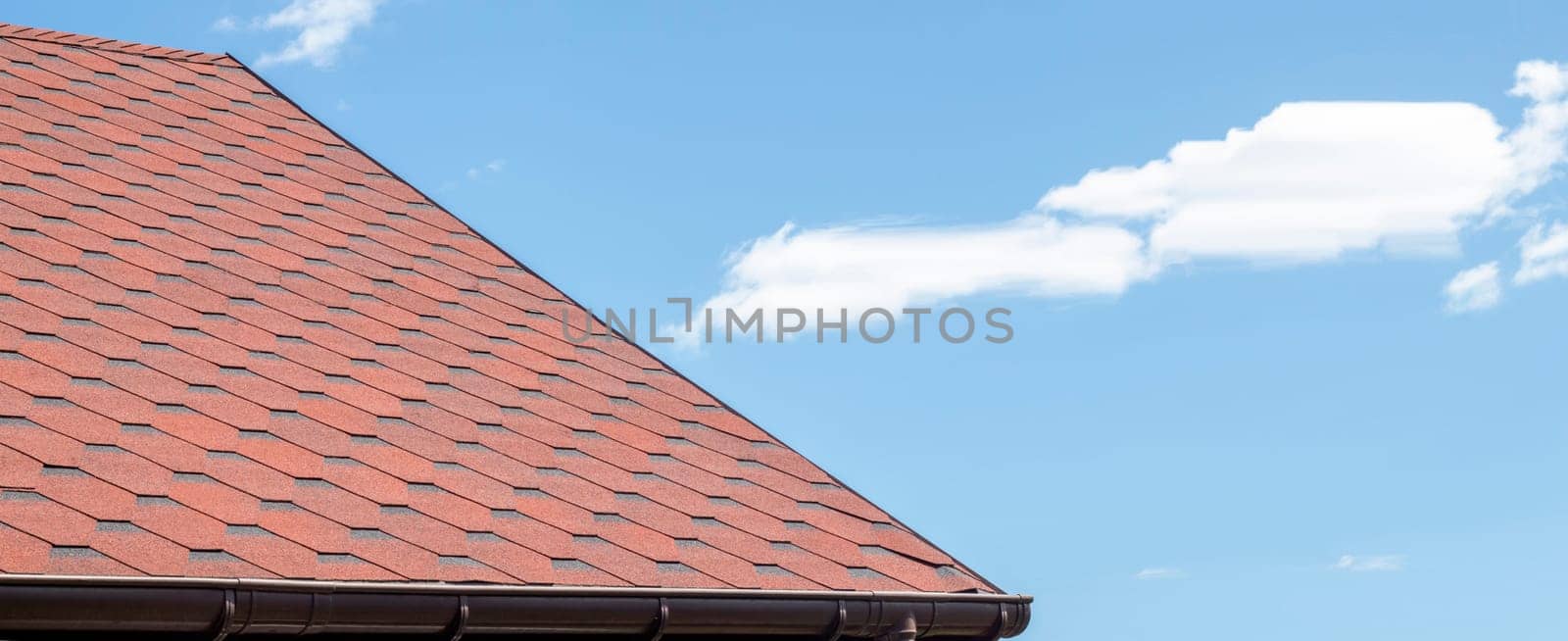 New roof with red shingles against the blue sky. Tiles on the roof of the house. Use to advertise roof fabrication and maintenance. Spotted texture. Affordable roofing. Banner with copy space
