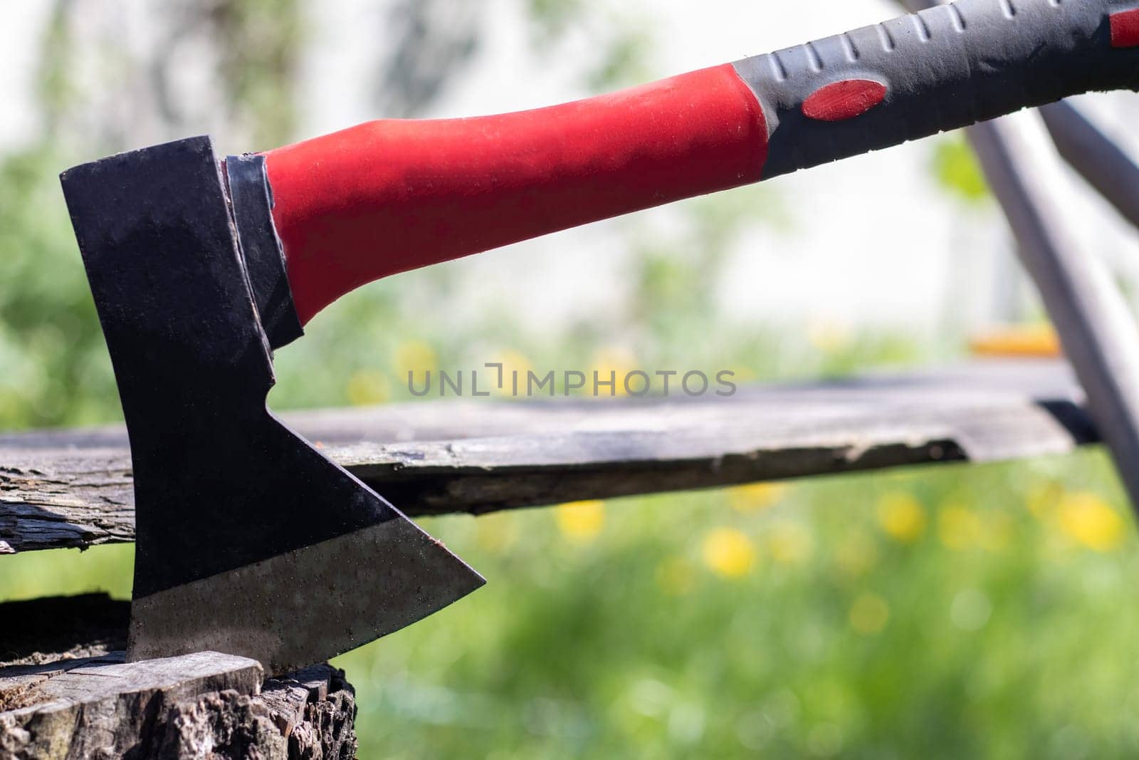 A new modern ax with a red handle sticks out of a wooden stump against a green meadow on a summer sunny day. The ax stuck into the stump. Ax blade in a log