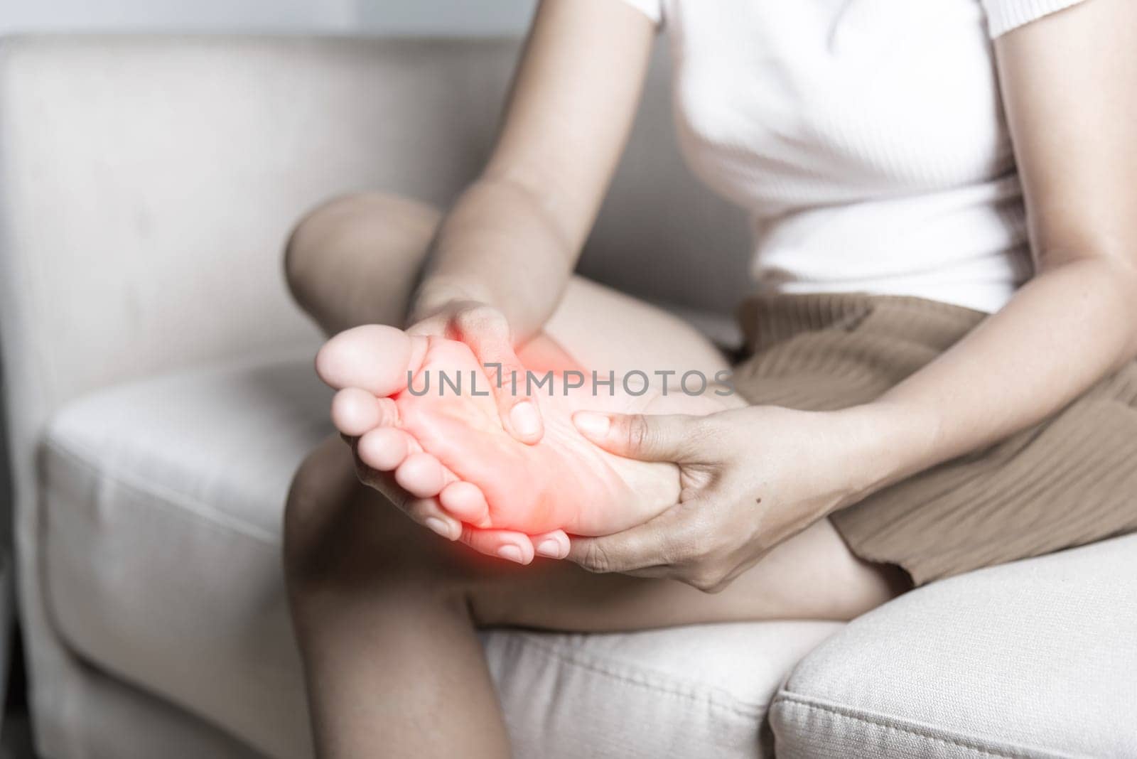 Foot pain, Asian woman sitting on sofa feeling pain in her foot at home, female suffering from feet ache use hand massage relax muscle from soles in home interior, Healthcare and podiatry medical