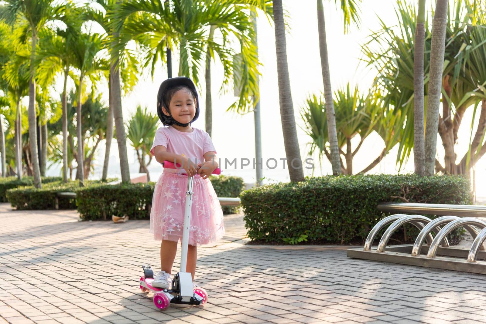 Child riding scooter. Happy Asian little kid girl wear safe helmet playing pink kick board on road in park outdoors on summer day, Active children games outside, Kids sport healthy lifestyle concept