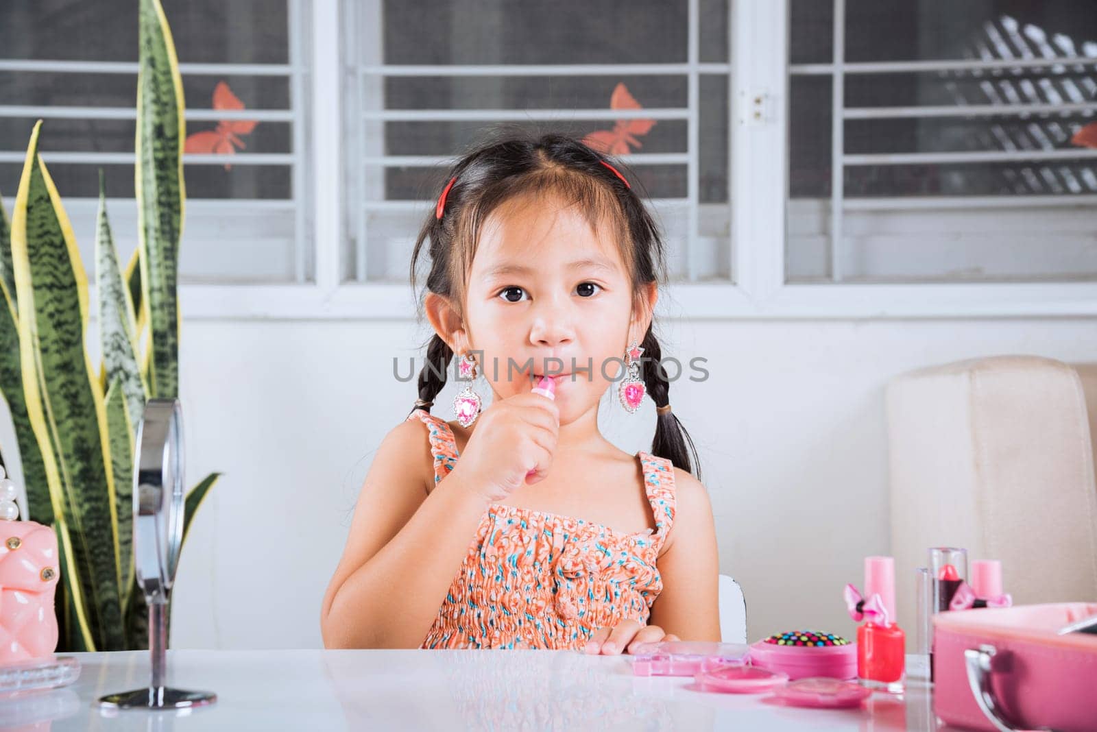 Asian adorable funny little girl making makeup her face she look in the mirror and applying red lipstick to mouth, Learning activity to be woman, happy kid is beautiful make up with cosmetics toy